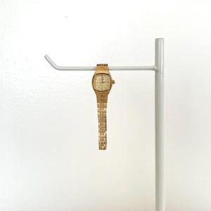 May include: A gold-tone analog wristwatch featuring a square-shaped case with rounded edges, a light champagne-colored dial with thin hour markers, and a textured link bracelet. The watch includes a small winding crown on the right side.