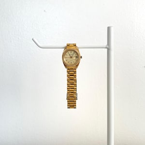 May include: A gold-toned wristwatch with a textured bezel and a matching link bracelet. The watch has a light-colored face with gold markers and hands. It is displayed on a white metal stand against a white background.