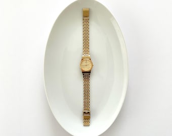 May include: A gold-tone analog wristwatch featuring a round face with a matching gold dial and a slender, intricate link bracelet. The watch is displayed vertically, showcasing its metallic finish and the delicate, repeating pattern of the band.