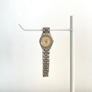 May include: A silver and gold-toned wristwatch with a light-colored face and a linked band. The watch is displayed on a white metal stand against a white background. The watch has a round face with small markers and hands.