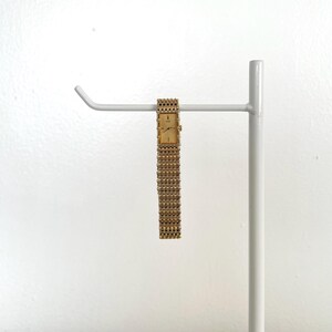 May include: A gold-toned rectangular wristwatch with a matching linked band hangs from a white metal display stand. The watch face is a light gold color, and the band is composed of small, closely connected links. The background is a plain white wall.