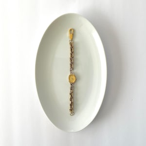 May include: A vintage gold-toned watch with a delicate chain band and a small, oval face. The watch is displayed on a white, oval-shaped plate, creating a minimalist aesthetic. The watch band is approximately 6 inches long.