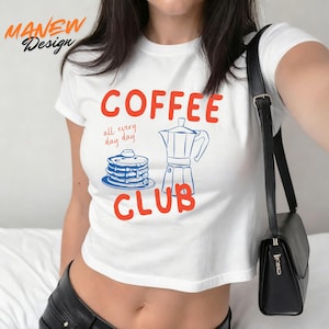 Op de afbeelding: Witte crop top met een rood en blauw ontwerp met de tekst "COFFEE CLUB" en een illustratie van pannenkoeken en een koffiezetapparaat. De tekst "all every day day" staat onder het woord "COFFEE". De merknaam "MANEW Design" staat linksboven.