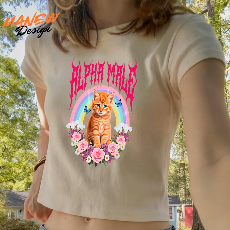 Pode incluir: Top cropped creme com um gr&aacute;fico de um gato malhado laranja, um arco-&iacute;ris e o texto "ALPHA MALE" em rosa. O gato &eacute; cercado por flores e borboletas. A parte superior tem mangas curtas.