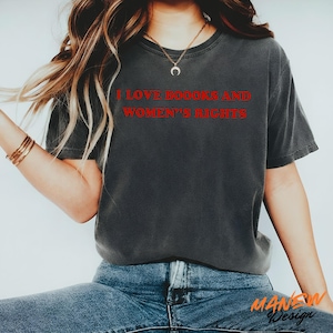 May include: A charcoal grey short-sleeved t-shirt featuring the red text I LOVE BOOOKS AND WOMEN'S RIGHTS printed on the chest. The garment has a crew neckline and a relaxed fit, paired with blue denim jeans and a gold crescent moon pendant necklace.