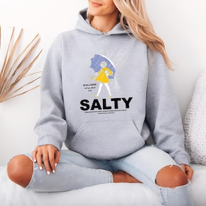 Puede incluir: Una sudadera con capucha gris claro con un gr&aacute;fico de una persona con impermeable amarillo sosteniendo un paraguas azul bajo la lluvia. La palabra "SALTY" est&aacute; impresa en letras negras grandes. La sudadera tiene un bolsillo delantero y una capucha con cord&oacute;n.