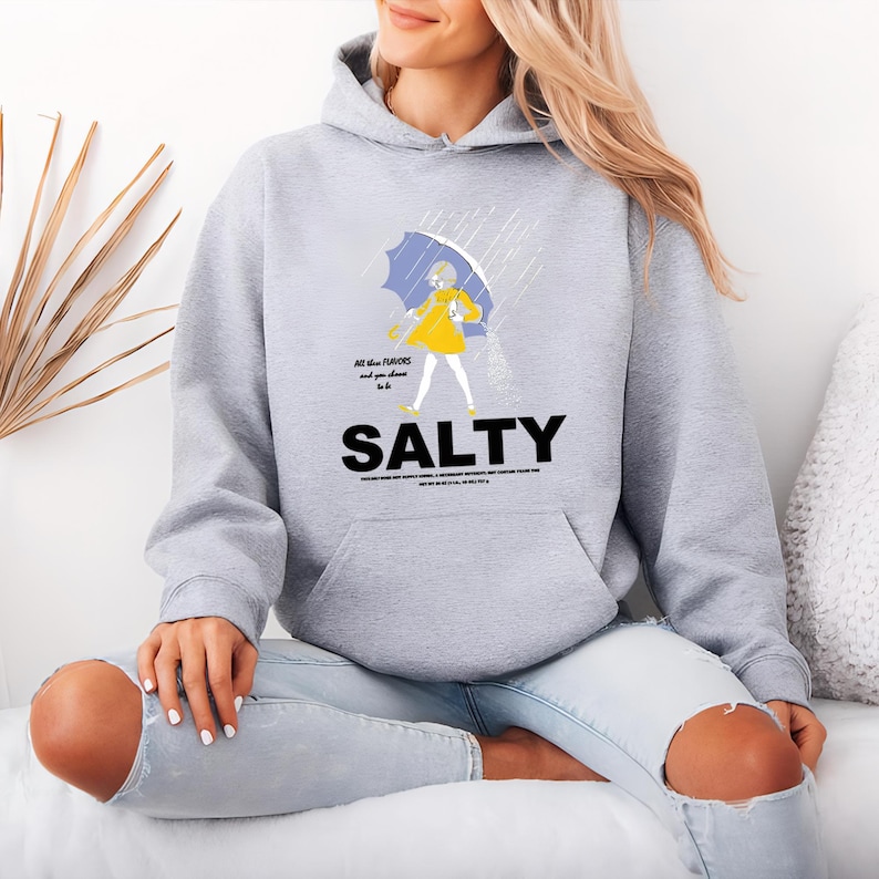 Puede incluir: Una sudadera con capucha gris claro con un gr&aacute;fico de una persona con impermeable amarillo sosteniendo un paraguas azul bajo la lluvia. La palabra "SALTY" est&aacute; impresa en letras negras grandes. La sudadera tiene un bolsillo delantero y una capucha con cord&oacute;n.