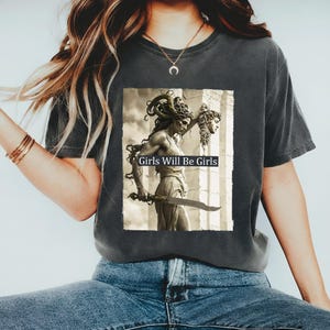 May include: Dark grey t-shirt with a sepia-toned graphic of a woman with Medusa-like hair holding a sword and a severed head. The text "Girls Will Be Girls" is printed below the image. A casual wear item.