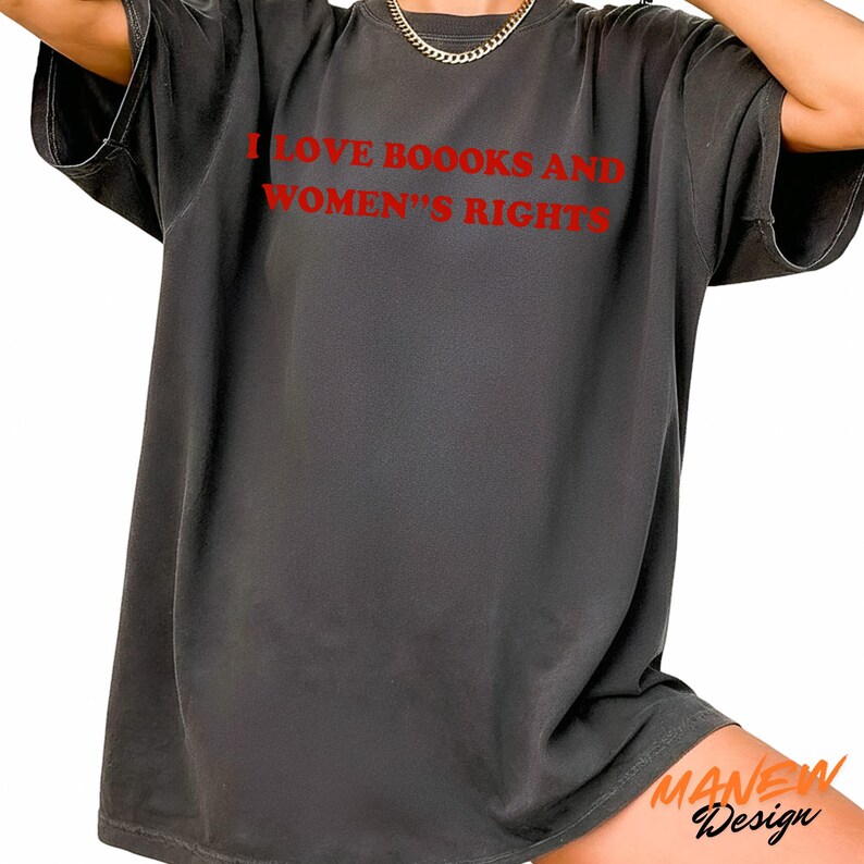 May include: A charcoal gray short-sleeve t-shirt featuring the red text I LOVE BOOOKS AND WOMEN'S RIGHTS printed on the chest. The garment has a crew neckline and a relaxed, loose-fitting silhouette with a slightly faded, washed-out fabric appearance.