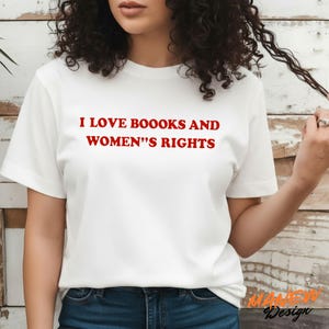 May include: A white short-sleeved t-shirt featuring the red text I LOVE BOOOKS AND WOMEN'S RIGHTS printed across the chest. The shirt has a crew neckline and a standard fit, providing a clean look with bold, centered lettering in a simple font.