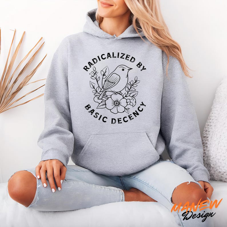 May include: A light gray hoodie with a front pocket and a drawstring hood. The graphic on the front features a bird and floral design with the text "RADICALIZED BY BASIC DECENCY" in a circular arrangement.