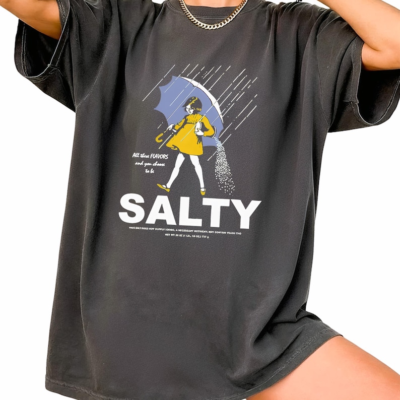 Puede incluir: Camiseta gris oscuro con un gr&aacute;fico de una persona bajo un paraguas azul, vertiendo sal. La palabra "SALTY" est&aacute; en letras blancas grandes debajo de la ilustraci&oacute;n. El texto encima de la ilustraci&oacute;n dice: "All these flavors and you choose to be".