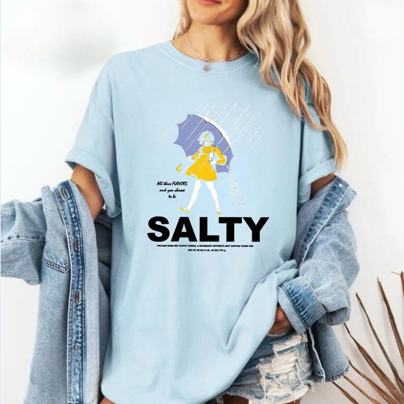 Puede incluir: Camiseta azul claro con la palabra "SALTY" en letras negras en negrita. La camiseta presenta un gr&aacute;fico de una persona bajo un paraguas bajo la lluvia, con el texto "All the flavors and you chose to be".