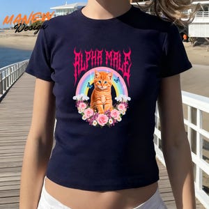 Pode incluir: Camiseta azul marinho com um gr&aacute;fico de um gato malhado laranja com olhos azuis, em frente a um arco-&iacute;ris e flores. O texto "ALPHA MALE" est&aacute; acima do gato em uma fonte rosa estilizada. A camiseta tem mangas curtas.