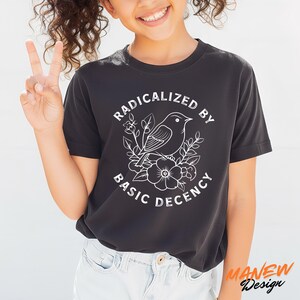 May include: Black t-shirt with a white graphic of a bird, flowers, and the text "RADICALIZED BY BASIC DECENCY." The shirt is a crew neck and has short sleeves. The brand name "MANEW Design" is printed in orange on the bottom right.