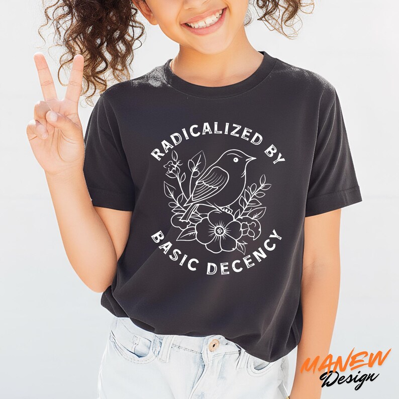 May include: Black t-shirt with a white graphic of a bird, flowers, and the text "RADICALIZED BY BASIC DECENCY." The shirt is a crew neck and has short sleeves. The brand name "MANEW Design" is printed in orange on the bottom right.