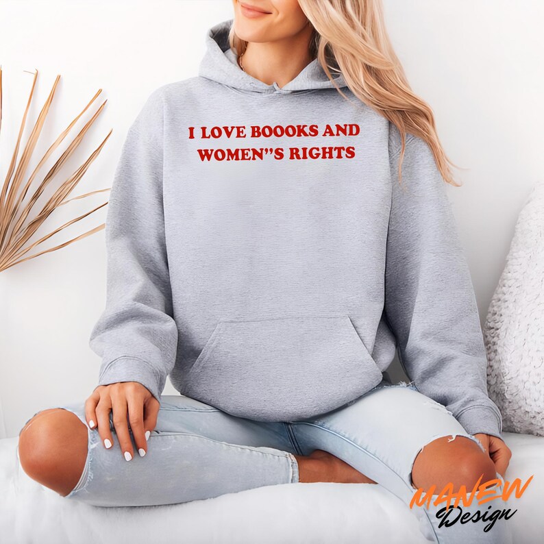 May include: A light gray hooded sweatshirt featuring the red text I LOVE BOOOKS AND WOMEN'S RIGHTS printed on the chest. The garment includes long sleeves, a front kangaroo pocket, and a hood with matching drawstrings, constructed from a soft fabric.