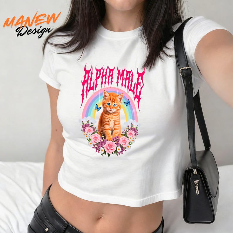 Pode incluir: Top curto branco com um gr&aacute;fico de um gato tigrado laranja, um arco-&iacute;ris e o texto "ALPHA MALE" em rosa. O design inclui rosas cor-de-rosa e borboletas azuis. Uma pe&ccedil;a de roupa casual.