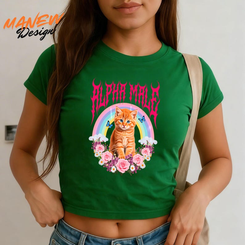 Pode incluir: Um top cropped verde com um design gr&aacute;fico. O design inclui um gato malhado laranja, um arco-&iacute;ris e o texto rosa "ALPHA MALE". O gato &eacute; cercado por flores e borboletas. O top tem mangas curtas.