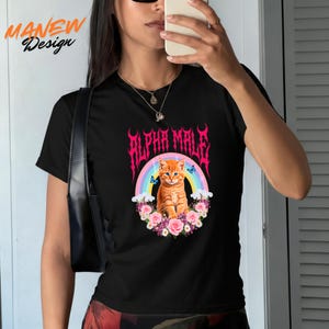 Pode incluir: Camiseta preta com um gr&aacute;fico de um gato tigrado laranja em frente a um arco-&iacute;ris, rodeado por flores e borboletas. O texto "ALPHA MALE" est&aacute; em rosa. O nome da marca "MANEW Design" est&aacute; no canto superior esquerdo.
