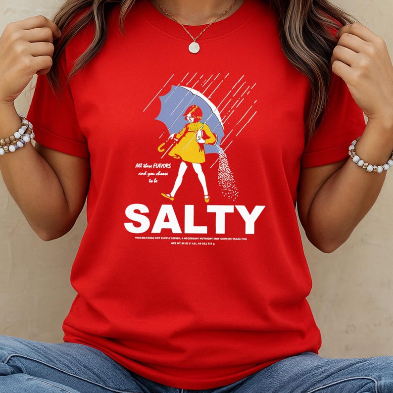 Puede incluir: Camiseta roja con un gr&aacute;fico de una persona bajo un paraguas azul, con sal cayendo. La palabra "SALTY" est&aacute; en letras blancas grandes debajo de la ilustraci&oacute;n, con la frase "All the flavors and you choose to be" arriba.