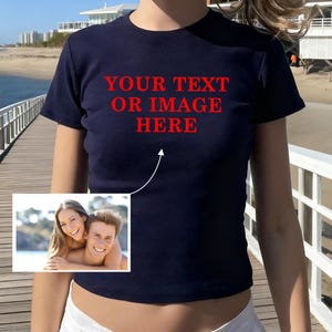 Può includere: T-shirt blu navy con la scritta rossa "YOUR TEXT OR IMAGE HERE". La maglietta presenta una piccola immagine di una coppia in basso a sinistra. La modella è in piedi su un molo di legno vicino alla spiaggia.