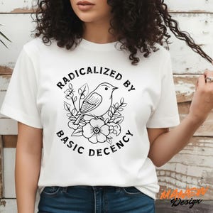 May include: White t-shirt featuring a black and white graphic of a bird perched on flowers, with the text "RADICALIZED BY BASIC DECENCY" in a circular design. The shirt is a classic crew neck style.