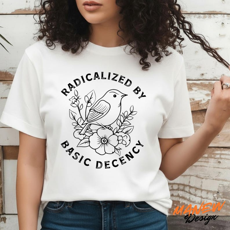 May include: White t-shirt featuring a black and white graphic of a bird perched on flowers, with the text "RADICALIZED BY BASIC DECENCY" in a circular design. The shirt is a classic crew neck style.