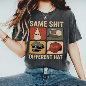 May include: Dark grey t-shirt featuring the text "SAME SHIT DIFFERENT HAT" above and below four images of hats. The hats include a white hood, a Confederate flag cap, a military hat, and a red "Make America Great Again" cap.