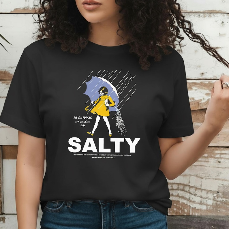 Puede incluir: Camiseta negra con un gr&aacute;fico de una persona bajo un paraguas azul, vertiendo sal. La persona lleva un vestido amarillo. El texto en la camiseta dice "All the flavors and you choose to be SALTY."