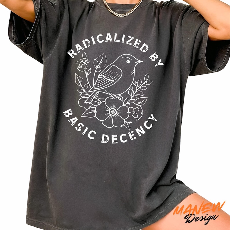 May include: Dark gray t-shirt with the text "RADICALIZED BY BASIC DECENCY" in a circular design. A white illustration of a bird and flowers is in the center. The shirt has short sleeves and a relaxed fit.