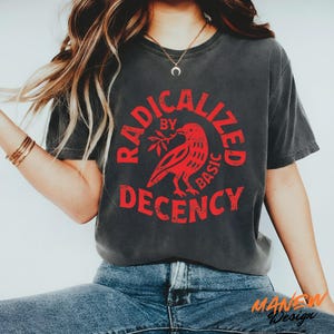 May include: A charcoal grey short-sleeve t-shirt featuring a red circular graphic with a bird holding a branch in the centre. The text RADICALIZED BY BASIC DECENCY is printed in a distressed red font around the bird illustration.