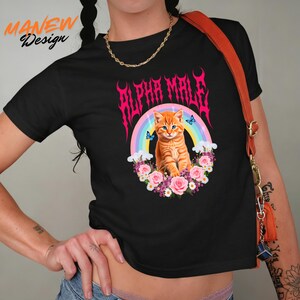 Pode incluir: Camiseta preta com um gr&aacute;fico de um gato laranja, um arco-&iacute;ris e o texto "ALPHA MALE" em rosa. O gato &eacute; cercado por flores e borboletas. A camiseta &eacute; um crop top.
