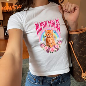 Pode incluir: Top curto branco com um gr&aacute;fico de um gato tigrado laranja, um arco-&iacute;ris e o texto rosa "ALPHA MALE". O gato &eacute; rodeado por flores e borboletas. A camisa tem mangas curtas.