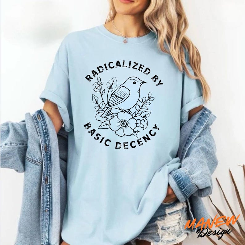 May include: Light blue t-shirt featuring a black and white illustration of a bird perched on flowers, with the text "RADICALIZED BY BASIC DECENCY" in a circular arrangement. The shirt has short sleeves and a relaxed fit.