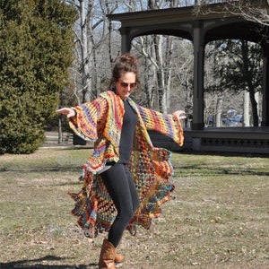 May include: A colourful, handmade crochet cardigan with long sleeves and an asymmetrical hemline. The open-front design displays a mix of orange, yellow, blue, and green hues. The garment's bohemian style is evident in its intricate crochet pattern.