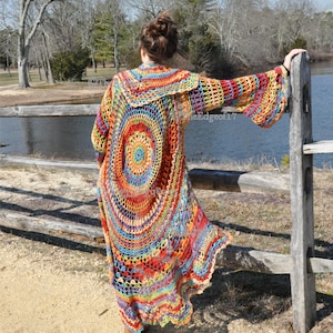 May include: A long, colourful crochet cardigan with a circular design on the back. The cardigan features a rainbow of colours, including red, orange, yellow, green, blue, and purple. The sleeves are bell-shaped. The person is standing near a wooden fence.