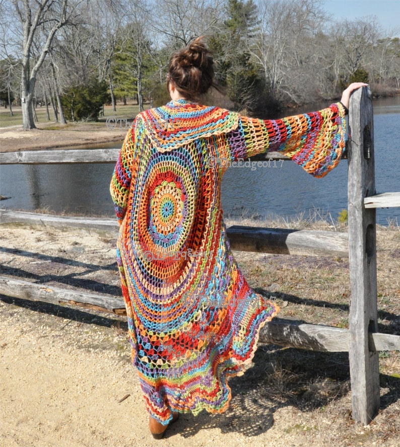 May include: A long, colourful crochet cardigan with a circular design on the back. The cardigan features a rainbow of colours, including red, orange, yellow, green, blue, and purple. The sleeves are bell-shaped. The person is standing near a wooden fence.