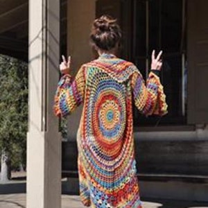 May include: A long, open-front crochet cardigan in a vibrant rainbow of colours. The back features a large circular design with concentric rings of red, orange, yellow, blue, and green. The sleeves are flared, and the cardigan is worn over a dark top and brown boots.