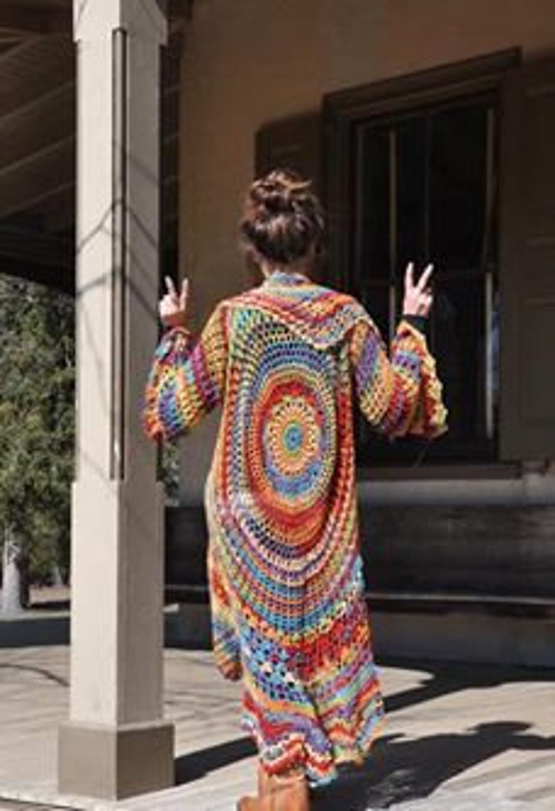 May include: A long, open-front crochet cardigan in a vibrant rainbow of colours. The back features a large circular design with concentric rings of red, orange, yellow, blue, and green. The sleeves are flared, and the cardigan is worn over a dark top and brown boots.