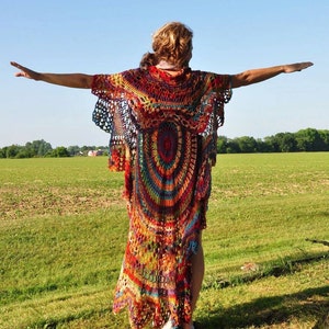 K&ouml;nnte beinhalten: Ein langer, bunter H&auml;kelcardigan mit einem kreisf&ouml;rmigen Muster in Rot-, Orange-, Gelb-, Gr&uuml;n-, Blau- und Violettt&ouml;nen. Der Cardigan wird von einer Person getragen, die auf einem Feld steht.