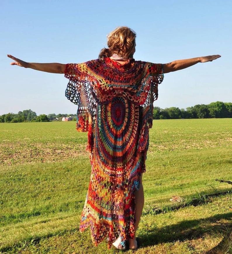 K&ouml;nnte beinhalten: Ein langer, bunter H&auml;kelcardigan mit einem kreisf&ouml;rmigen Muster in Rot-, Orange-, Gelb-, Gr&uuml;n-, Blau- und Violettt&ouml;nen. Der Cardigan wird von einer Person getragen, die auf einem Feld steht.