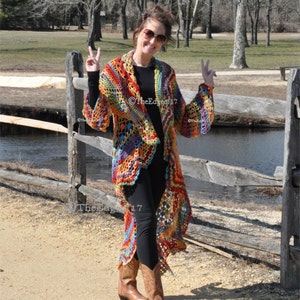 May include: A long, open-front crochet cardigan in a vibrant rainbow of colours. The handmade garment features a loose, flowing design with a textured stitch pattern. The cardigan is worn with black leggings and brown cowboy boots. The Edge of 17 is printed on the bottom left.