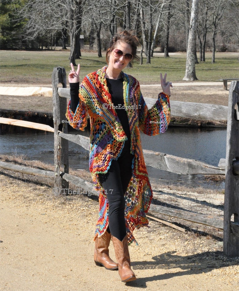 May include: A long, open-front crochet cardigan in a vibrant rainbow of colours. The handmade garment features a loose, flowing design with a textured stitch pattern. The cardigan is worn with black leggings and brown cowboy boots. The Edge of 17 is printed on the bottom left.