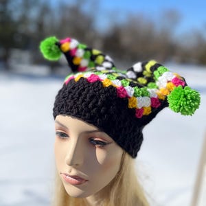May include: A black crochet hat with a colorful patchwork design and two pointed tips, each topped with a bright green pom-pom. The hat features a mix of pink, yellow, white, and green squares.