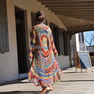 May include: A long, open-front crochet cardigan in a vibrant rainbow of colours. The design features a large circular pattern on the back, with long sleeves and a flowing, floor-length hem. The Edge logo is visible on the garment.