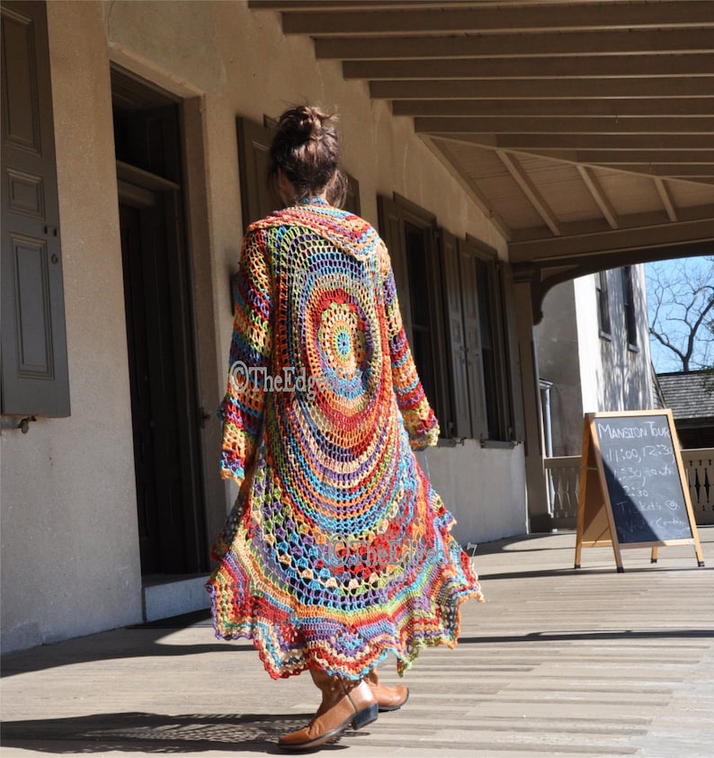May include: A long, open-front crochet cardigan in a vibrant rainbow of colours. The design features a large circular pattern on the back, with long sleeves and a flowing, floor-length hem. The Edge logo is visible on the garment.