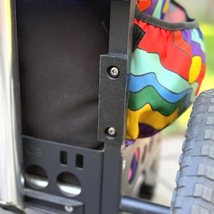 Puede incluir: Primer plano de un carrito utilitario negro y plateado. El carrito tiene un marco negro, un compartimento de tela negra y un bolsillo lateral estampado de colores. Tiene una parte superior acolchada azul y un neum&aacute;tico negro.