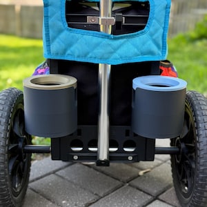 Puede incluir: Un carrito utilitario negro y plateado con dos ruedas negras y dos portavasos grises. El carrito tiene una cubierta acolchada azul y un asa plateada. El carrito est&aacute; dise&ntilde;ado para uso en exteriores.