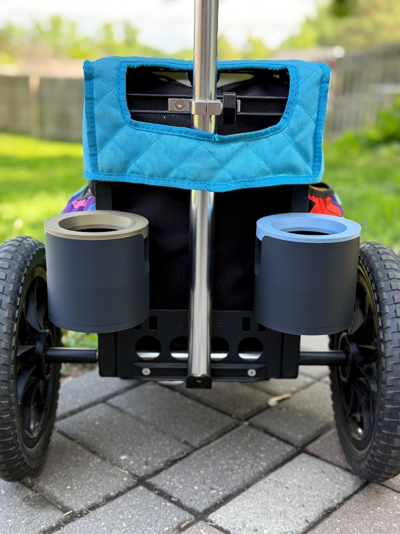 Puede incluir: Un carrito utilitario negro y plateado con dos ruedas negras y dos portavasos grises. El carrito tiene una cubierta acolchada azul y un asa plateada. El carrito est&aacute; dise&ntilde;ado para uso en exteriores.